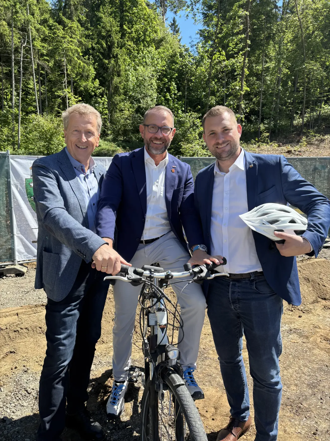 Three men in suits and glasses stand next to a bicycle. One man holds a helmet. They appear to be posing for a photograph at a construction site. Trees and a fence are in the background.