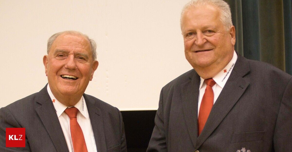 Zwei ältere Männer, beide in Anzügen und roten Krawatten, lächeln für ein Foto vor einem weißen Hintergrund.