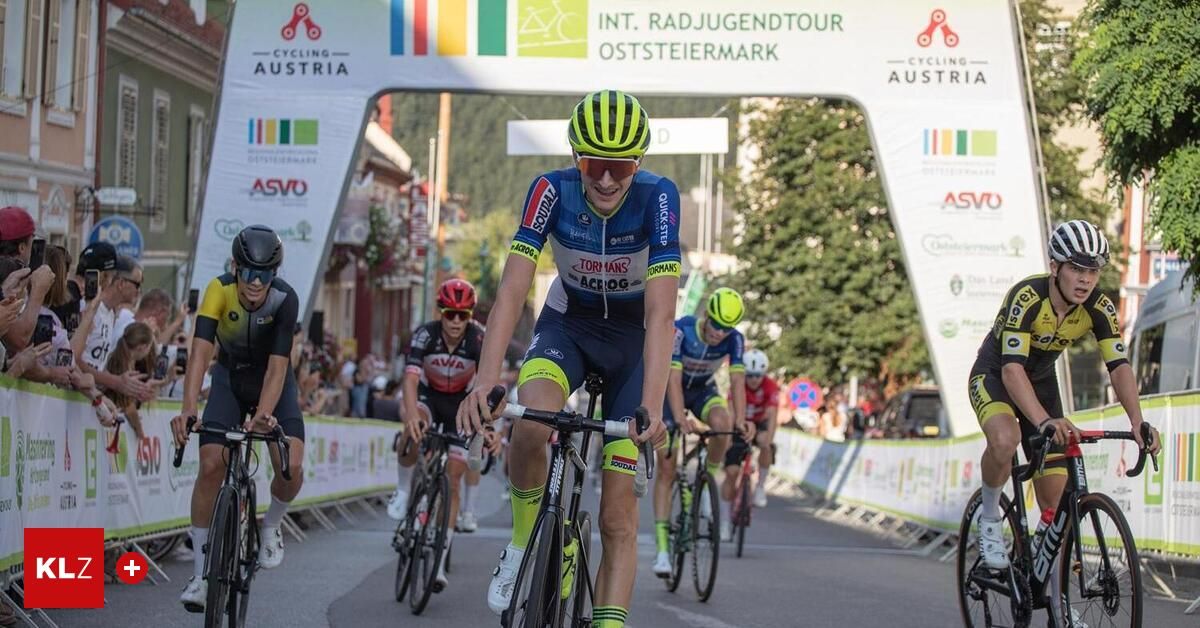 Eine Gruppe von Radfahrern fährt auf einer Straße, mit einem Banner über ihnen. Sie tragen Helme und Radfahreranzüge. Der Mann in der Mitte lächelt und scheint das Rennen zu führen. Hinter ihm befinden sich andere Radfahrer, die ebenfalls Rennen fahren.