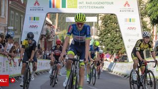 Eine Gruppe von Radfahrern fährt auf einer Straße, mit einem Banner über ihnen. Sie tragen Helme und Radfahreranzüge. Der Mann in der Mitte lächelt und scheint das Rennen zu führen. Hinter ihm befinden sich andere Radfahrer, die ebenfalls Rennen fahren.