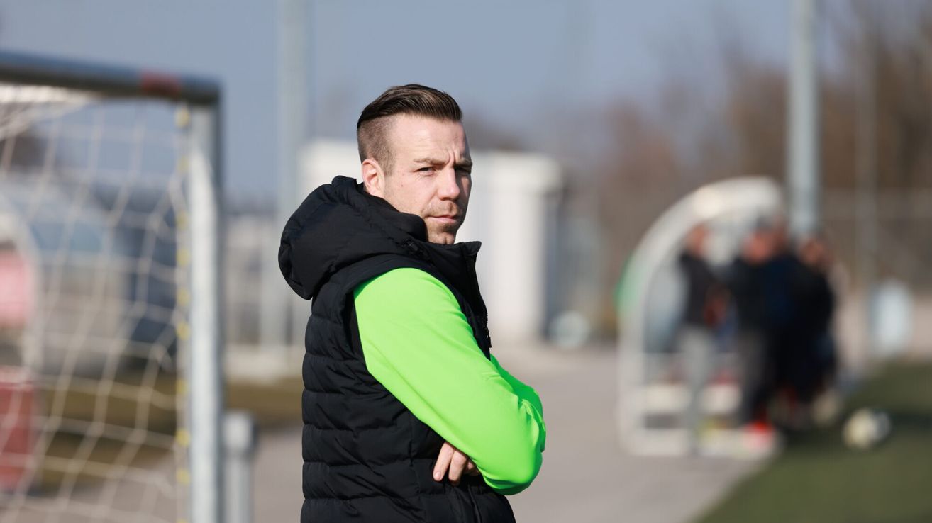A man with a serious expression stands on a field, wearing a black puffer vest with a green sleeve. His arms are crossed, and he looks ahead. Behind him, there are blurred images of buildings and people.