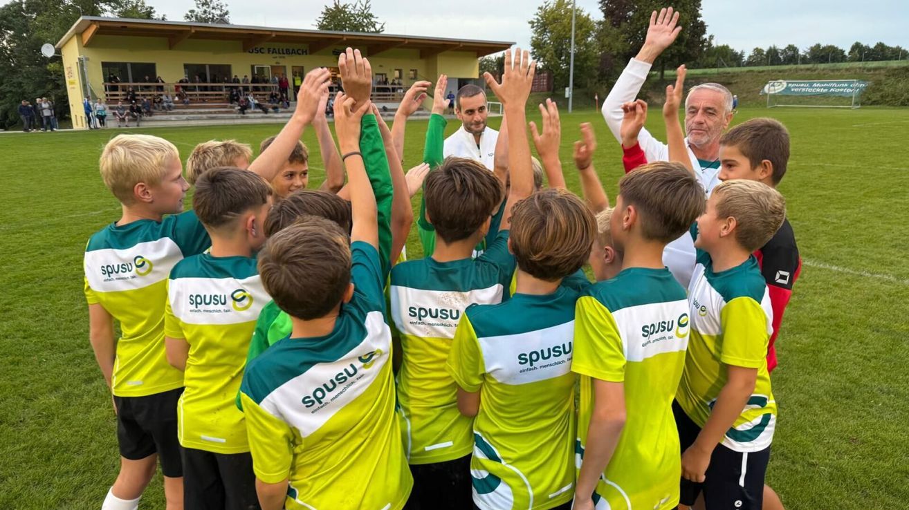 Eine Gruppe junger Jungen in grünen und gelben Trikots hebt auf einem Fußballfeld die Hände. Ein erwachsener Mann steht hinter ihnen, und eine andere Person ist neben ihm. Im Hintergrund sitzen Leute auf Tribünen.