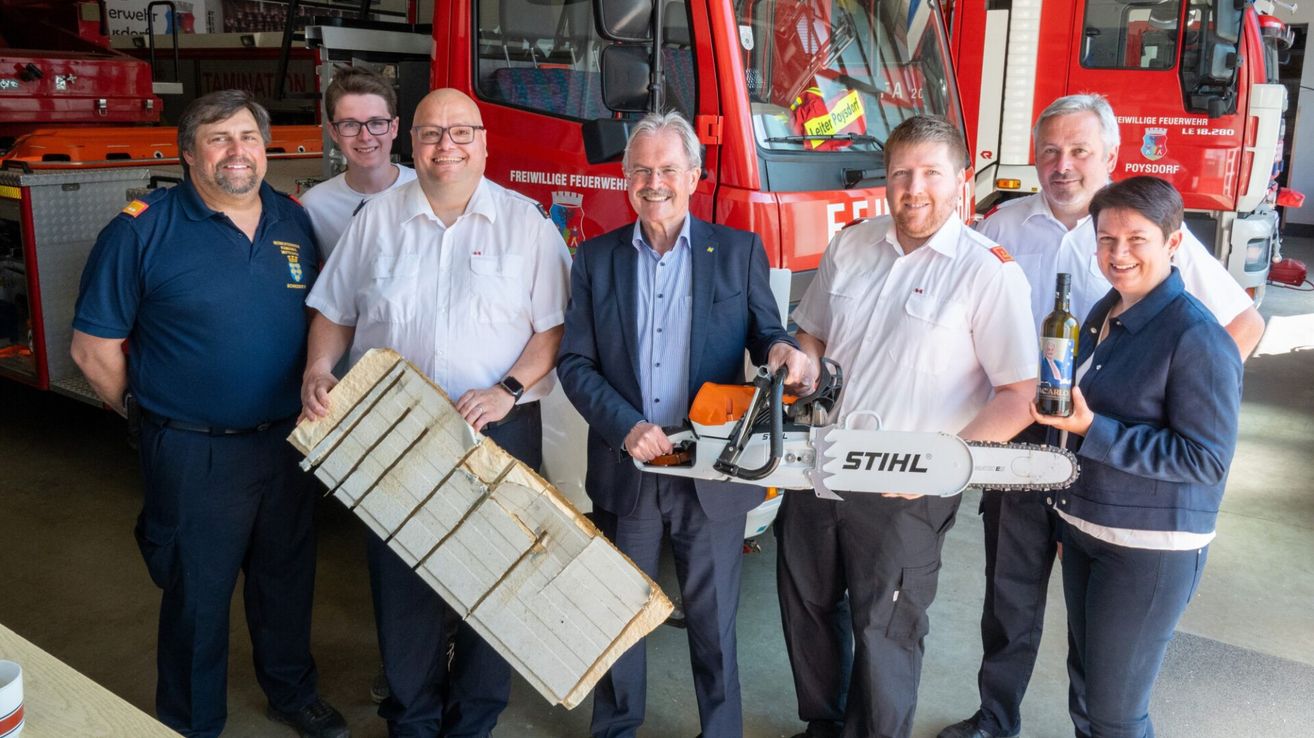 Fünf Männer stehen vor einem roten Feuerwehrauto und lächeln. Der Mann in der Mitte hält eine Kettensäge, während der auf der linken Seite ein Holzbrett hält.