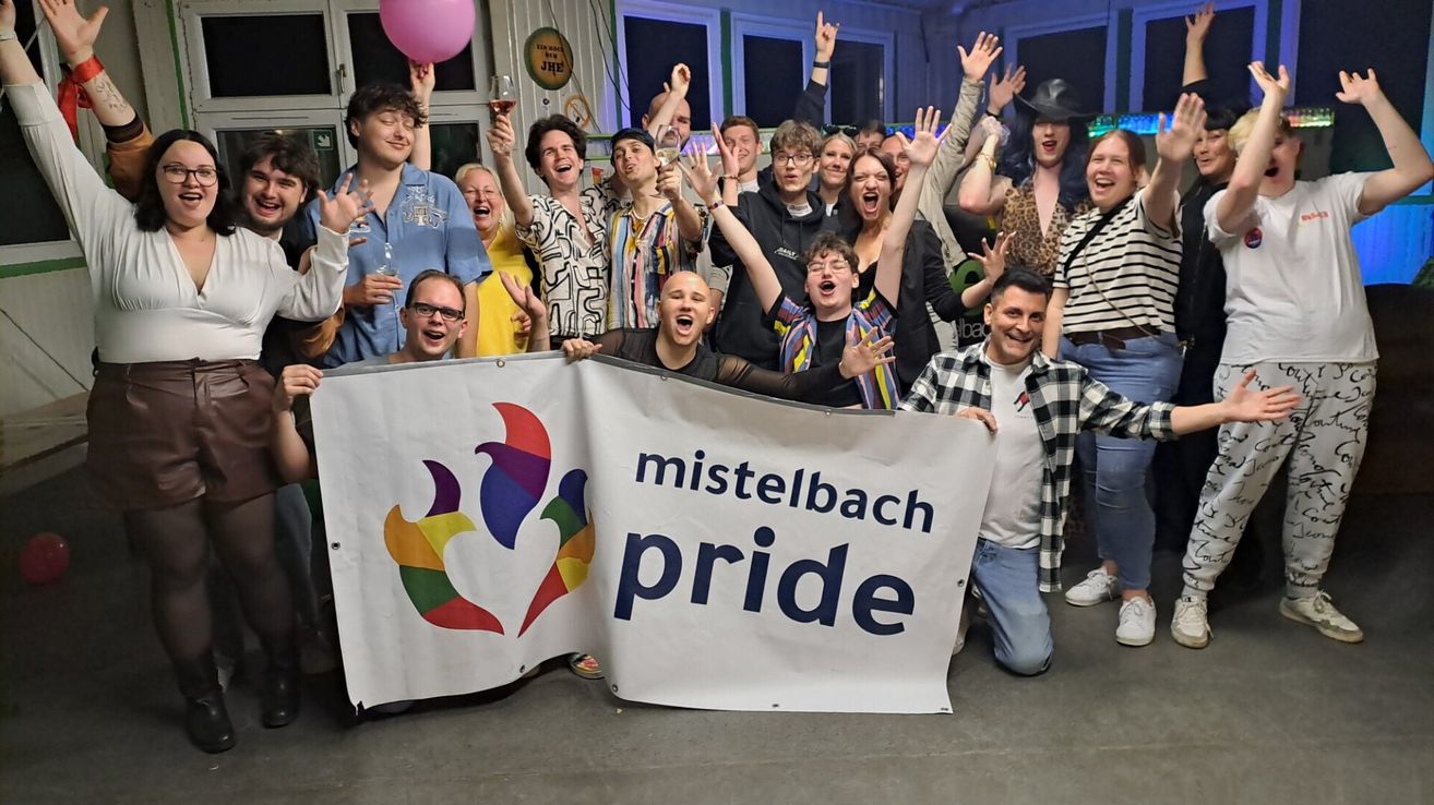 Eine Gruppe von Menschen feiert mit einem Banner, auf dem 'mistelbach pride' steht. Sie lächeln und halten ihre Hände und Getränke hoch.