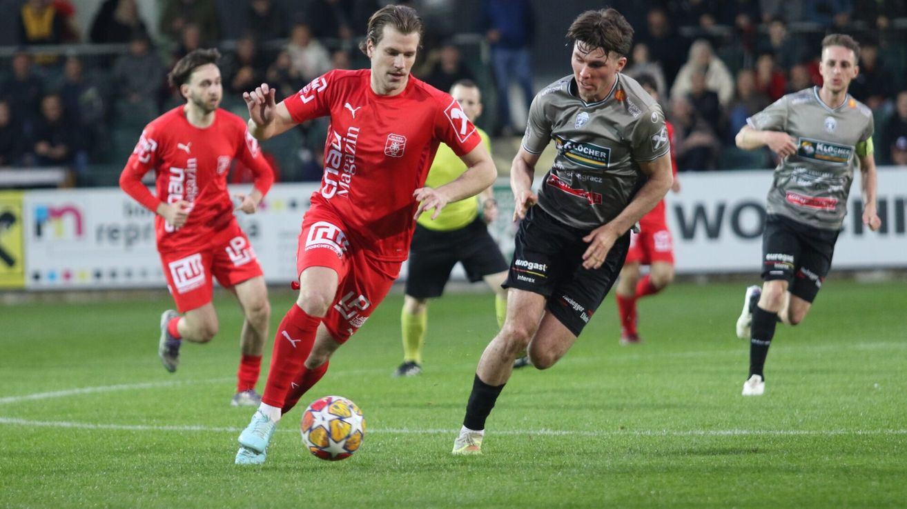Ein Fußballspiel ist im Gange mit Spielern in roten und grauen Trikots. Ein Spieler in Rot dribbelt den Ball, während ein anderer in Grau versucht, ihn zu stehlen. Zuschauer beobachten im Hintergrund.
