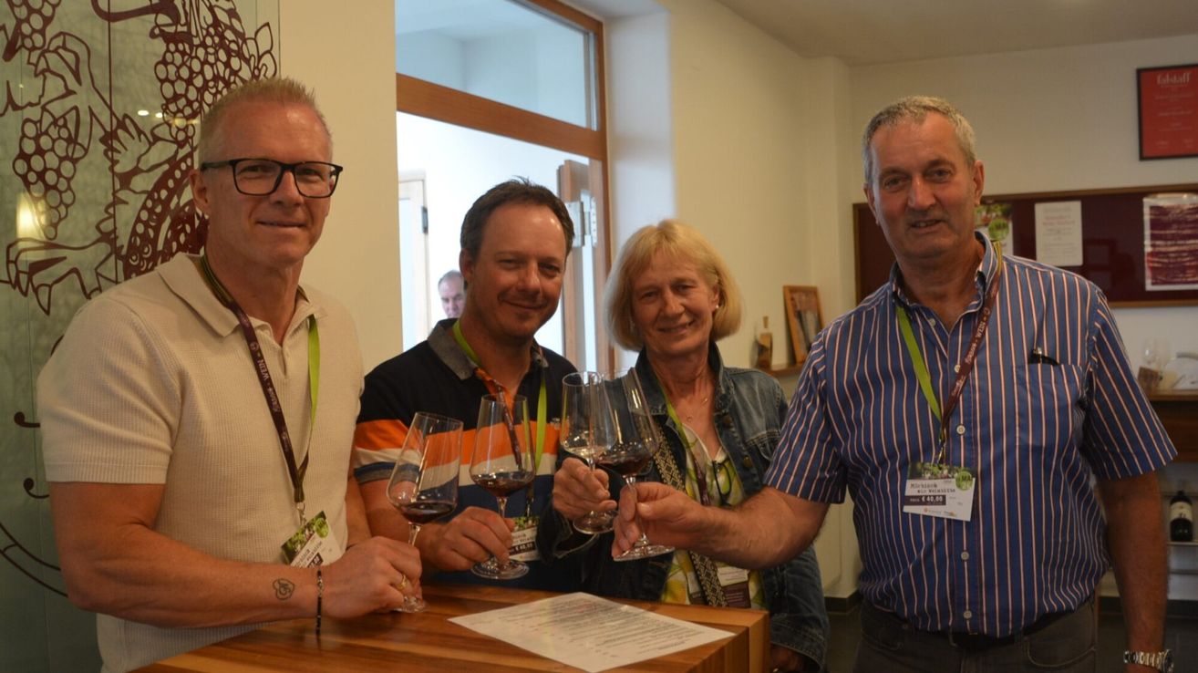 Vier Personen stehen an einem Tresen und halten Weingläser mit Wein gefüllt in der Hand. Sie lächeln und scheinen für ein Foto zu posieren.