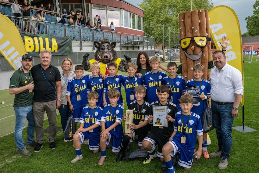 Eine Fußballmannschaft posiert mit Trainern und einem Maskottchen vor einem Stadion. Sie halten Medaillen und Trophäen, mit einer Menge im Hintergrund.