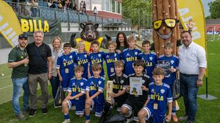 Eine Fußballmannschaft posiert mit Trainern und einem Maskottchen vor einem Stadion. Sie halten Medaillen und Trophäen, mit einer Menge im Hintergrund.