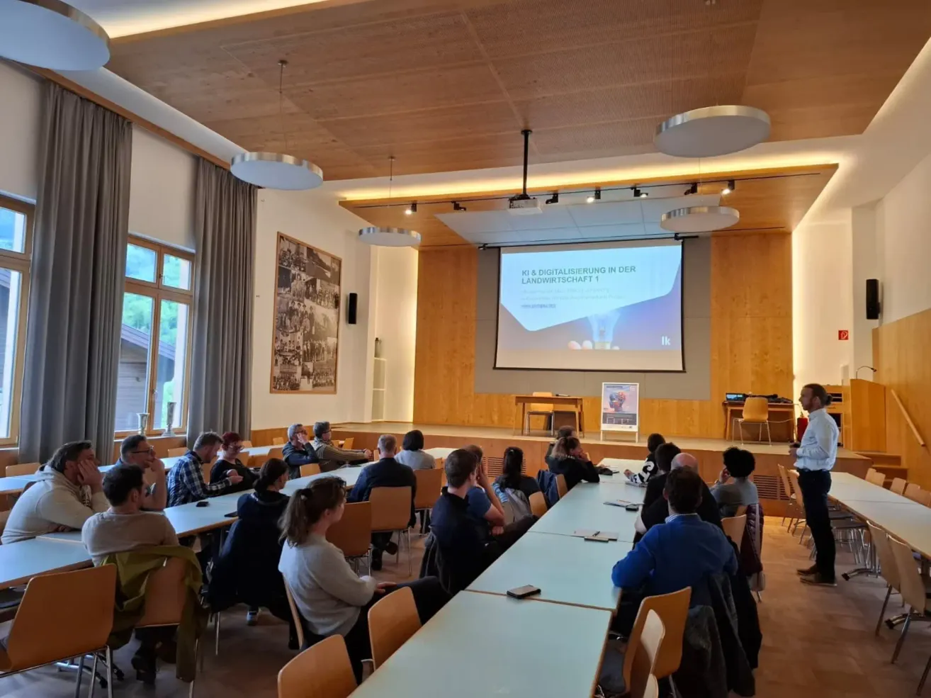Ein Raum mit einer Projektionsleinwand, auf der eine Präsentation angezeigt wird. Die Leute sitzen an langen Tischen und sind auf die Präsentation konzentriert. Es gibt Holzwände und Deckenleuchten.