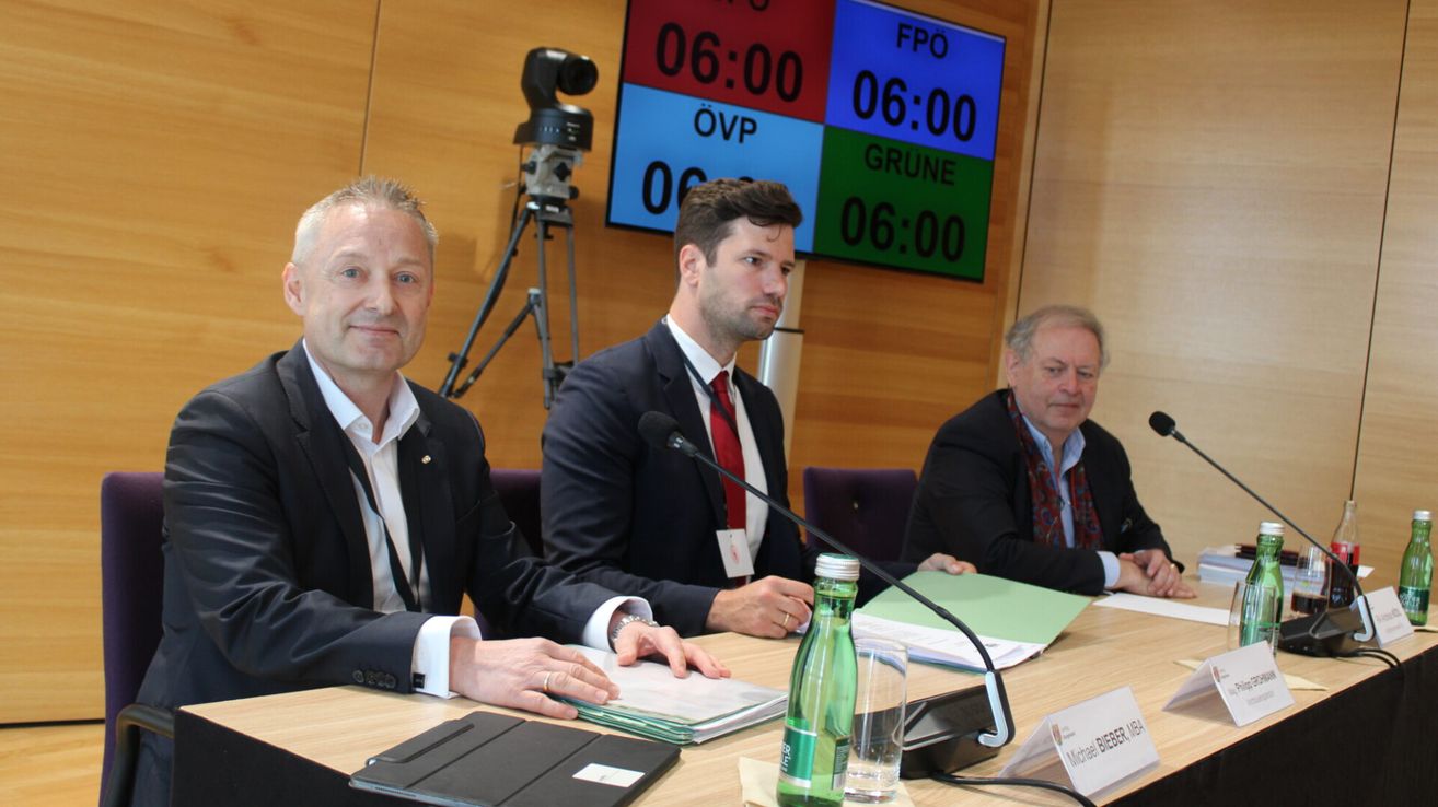 Drei Männer in Anzügen sitzen an einem Konferenztisch mit Mikrofonen und Papieren vor sich. Ein digitaler Timer zeigt '06:00' in drei Farben an einer Wand hinter ihnen.