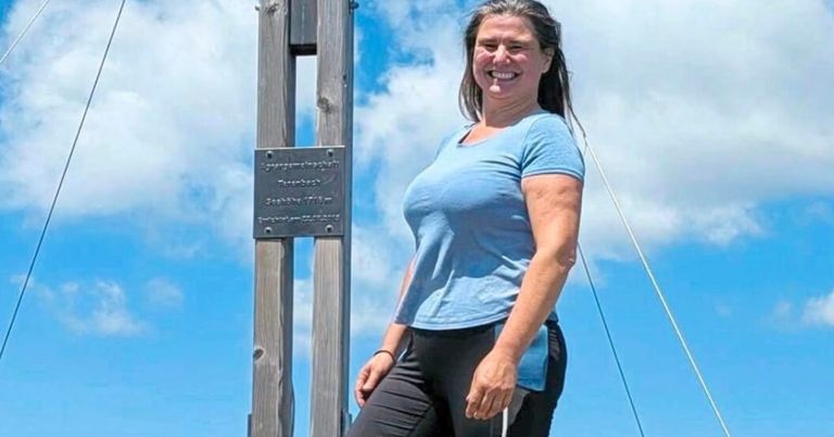 Eine Frau in einem blauen T-Shirt und schwarzen Hosen steht lächelnd neben einer Holzkonstruktion mit einem Schild und einer Metallplatte.