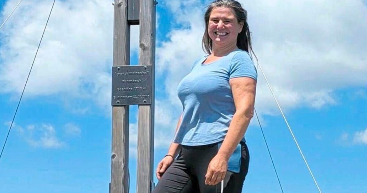 Eine Frau in einem blauen T-Shirt und schwarzen Hosen steht lächelnd neben einer Holzkonstruktion mit einem Schild und einer Metallplatte.