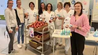 Eine Gruppe von Frauen in weißen Uniformen und Brillen steht neben einem Tisch mit Äpfeln und Reinigungsprodukten. Eine Frau in einem rosa Pullover hält ein Buch und lächelt.
