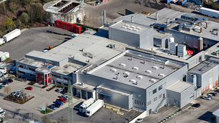 Luftaufnahme eines Industriekomplexes mit großen Lagerhäusern, einem roten Lagergebäude und mehreren vor dem Gebäude geparkten Fahrzeugen.