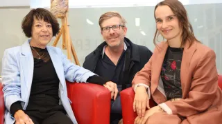 Drei Personen, zwei Frauen und ein Mann, sitzen auf roten Stühlen. Die Frau auf der linken Seite trägt eine hellblaue Jacke und die Frau auf der rechten Seite trägt einen pfirsichfarbenen Mantel. Der Mann trägt eine Brille.
