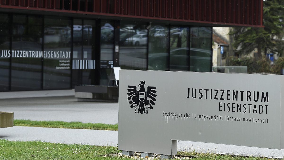 Ein Schild für Justizzentrum Eis mit einem deutschen Adler-Emblem steht vor einem Gerichtsgebäude. Hinter dem Schild befinden sich Glaswände mit Reflexionen von Autos und Gebäuden.