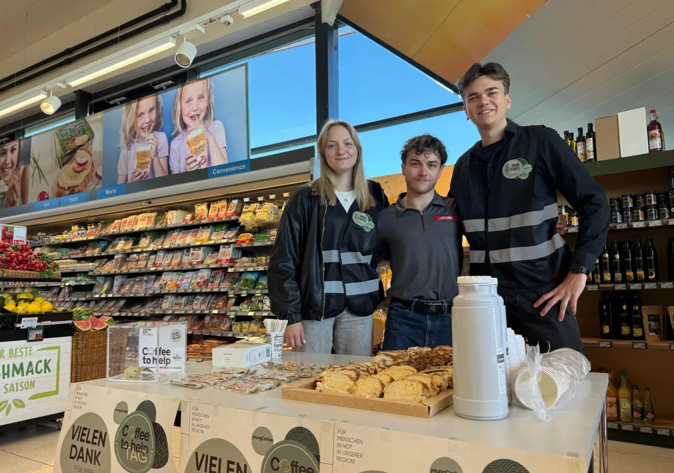 Three young people, likely volunteers, stand behind a table in a convenience store, smiling and posing for a photo. A sign on the table reads 'Coffee to Help'. Various foods are displayed on the table and shelves behind them.
