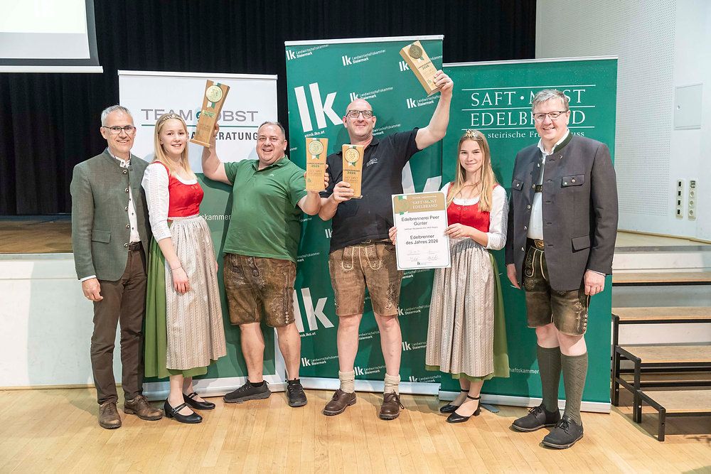 Fünf Personen, vier mit Auszeichnungen, stehen auf einer Bühne vor einem grünen Hintergrund. Sie tragen traditionelle Kleidung. Der Mann rechts trägt einen Anzug und Lederschuhe. Die Frau links hält eine Urkunde.