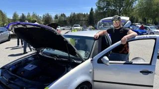 Ein Mann steht neben einem silbernen Auto mit geöffneter Motorhaube, die den Motor zeigt. Im Hintergrund sind Bäume und geparkte Autos zu sehen.