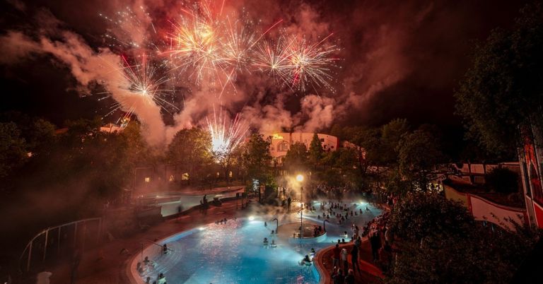 A fireworks display at night over a pool with people swimming and standing around.