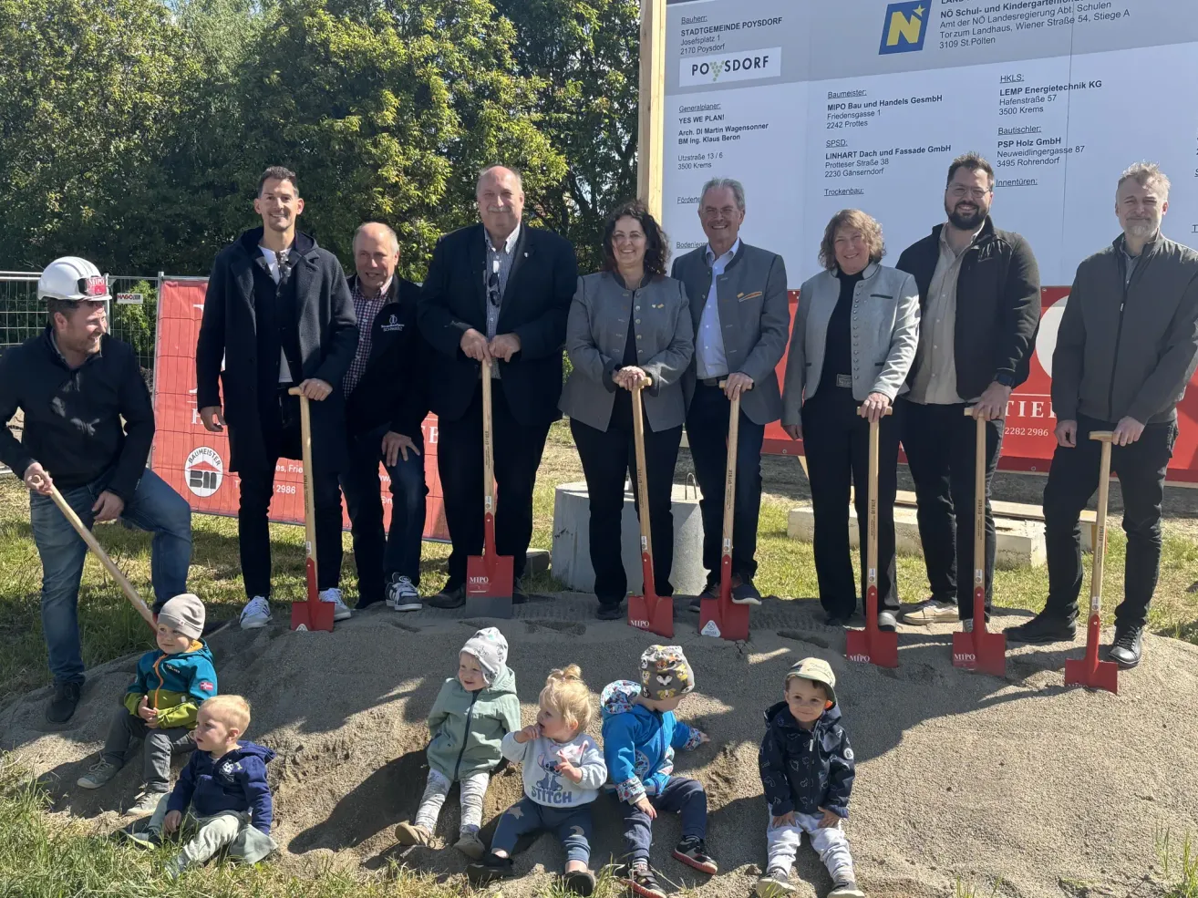Mehrere Erwachsene und Kinder posieren mit Schaufeln auf einer Baustelle für ein Foto. Hinter ihnen steht ein großes Schild mit der Aufschrift 'POSDORF'. Im Hintergrund sind Bäume und ein Zaun zu sehen.