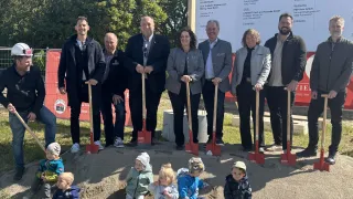 Mehrere Erwachsene und Kinder posieren mit Schaufeln auf einer Baustelle für ein Foto. Hinter ihnen steht ein großes Schild mit der Aufschrift 'POSDORF'. Im Hintergrund sind Bäume und ein Zaun zu sehen.