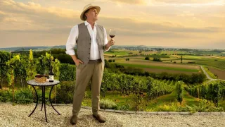 Ein Mann in einer Weste steht auf einer Terrasse und hält ein Weinglas, vor ihm ein Weinberg.