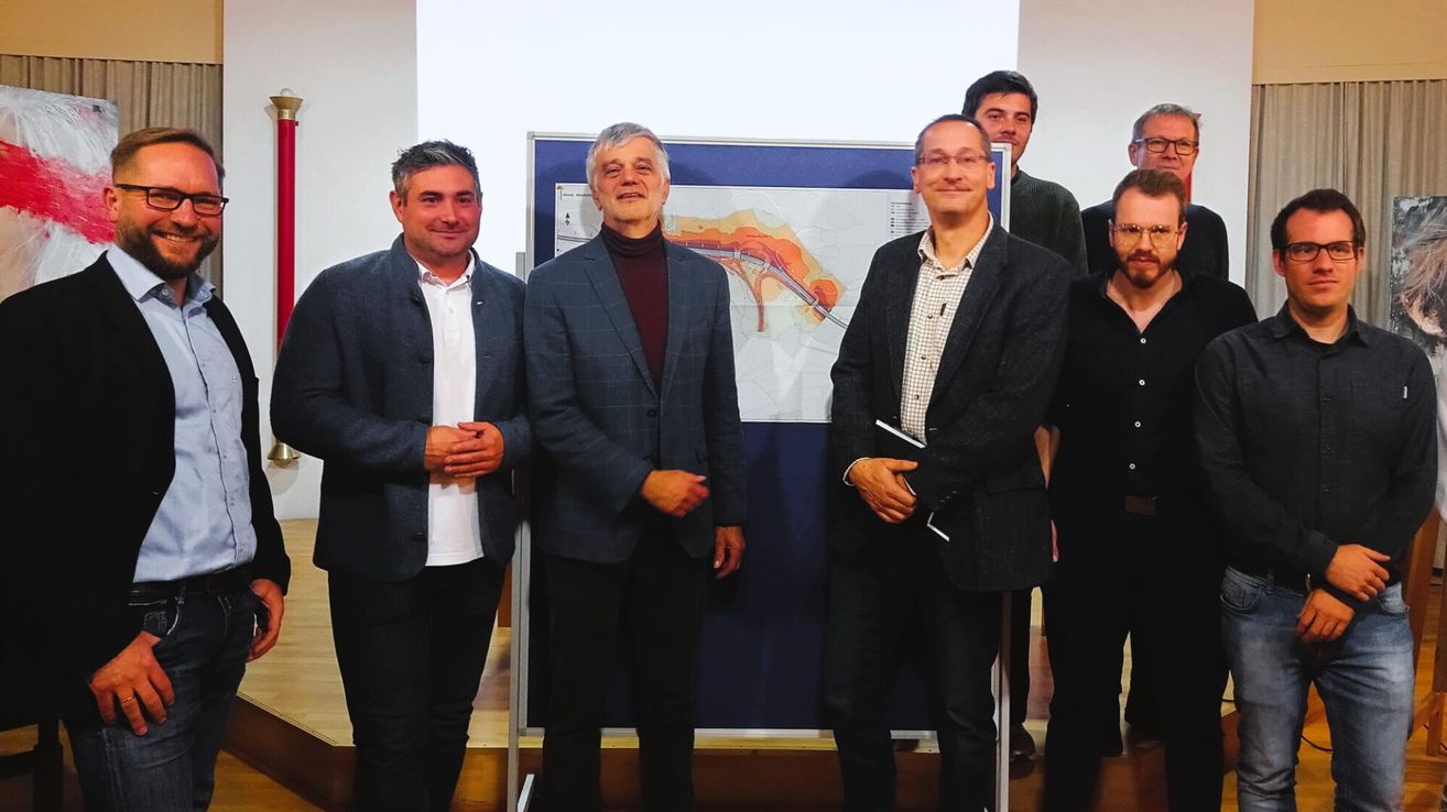 Five men stand in front of a large poster. The central man holds a folder. Two men on the sides smile. Another man behind them looks at the camera.