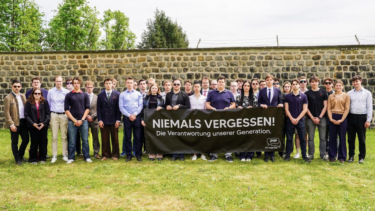 A group of individuals stands behind a black banner with white German text. They are dressed in formal attire. Behind them is a stone wall with barbed wire.