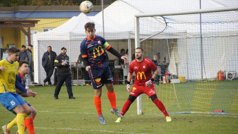 Zwei Fußballspieler in Aktion auf dem Feld. Ein Spieler springt, um den Ball zu köpfen, während der andere das Tor verteidigt. Dahinter befinden sich Zuschauer und ein Tisch mit Erfrischungen.