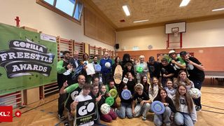 Eine Gruppe von Menschen in einer Turnhalle mit einem Basketballkorb. Sie halten ein Frisbee, einen Ball und ein Zertifikat. Einige von ihnen sitzen auf dem Boden, während andere stehen. Der Raum hat einen Holzboden und eine weiße Wand mit einer Uhr und einem Plakat.