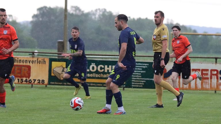 Drei Männer spielen Fußball auf einem Feld im Regen. Ein Mann trägt Blau, ein anderer Gelb und der dritte Rot. Sie schauen auf einen Fußball auf dem Boden.