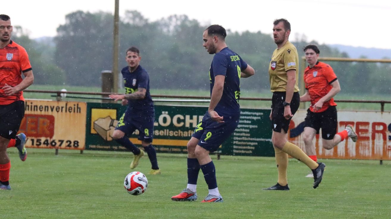 Drei Männer spielen Fußball auf einem Feld im Regen. Ein Mann trägt Blau, ein anderer Gelb und der dritte Rot. Sie schauen auf einen Fußball auf dem Boden.
