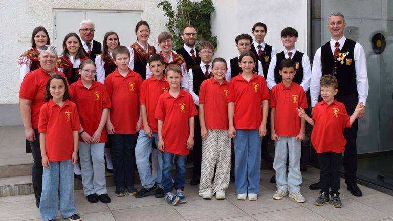 A group of children and adults in red polo shirts are posing for a photo outside. They stand in two rows, with a few adults behind them.