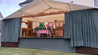 Vier Personen sitzen an einem Tisch mit roter Tischdecke unter einem Baldachin und probieren wahrscheinlich für eine Theateraufführung. Die Bühne ist von blauen Vorhängen umrahmt.