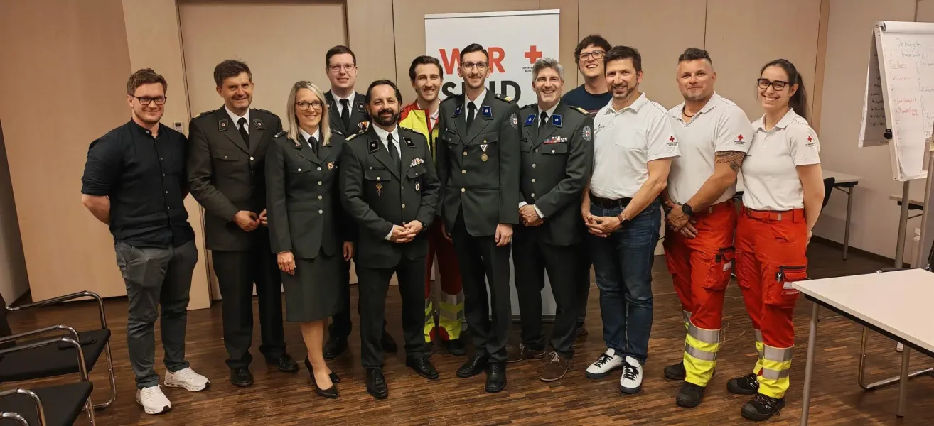 Eine Gruppe uniformierter Personen, darunter sowohl Männer als auch Frauen, stehen gemeinsam vor einem Schild mit dem Roten Kreuz Logo.