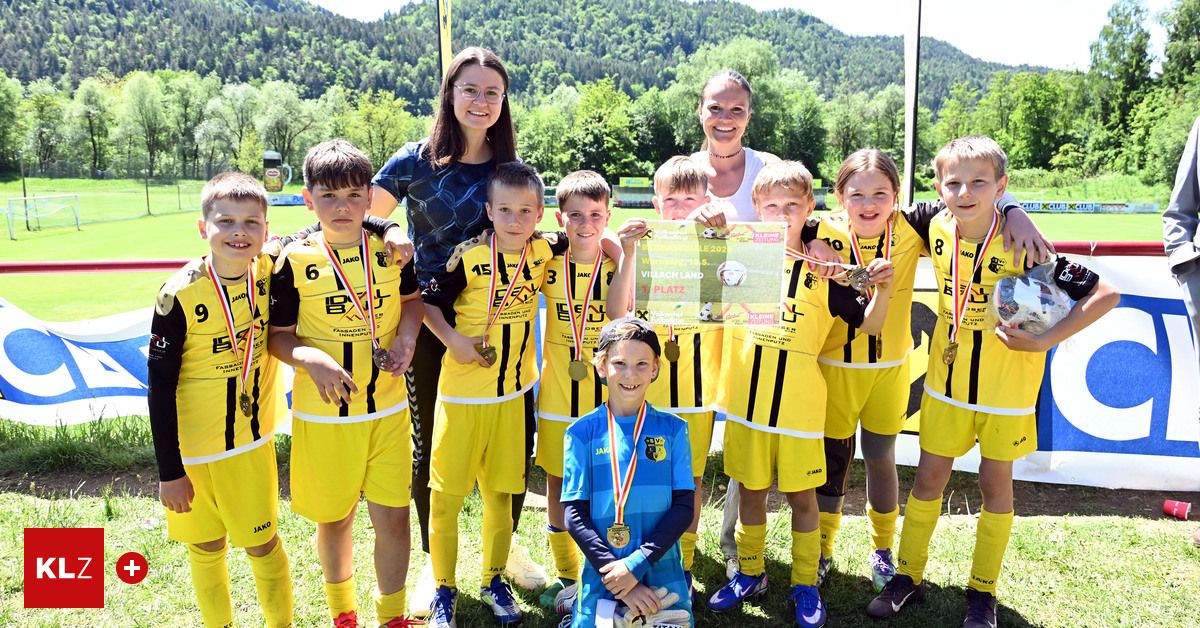 Eine Gruppe von Kindern und Erwachsenen in gelben und schwarzen Uniformen, wahrscheinlich eine Fußballmannschaft, posiert mit ihren Medaillen auf einem Rasenfeld. Zwei Frauen lächeln und stehen hinter ihnen.