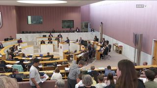 Eine Parlamentssitzung ist im Gange, die Teilnehmer sitzen im Halbkreis. Eine zentrale Figur steht an einem Rednerpult, und Monitore sind an den Wänden montiert. Zuschauer sitzen im Vordergrund und beobachten die Vorgänge.