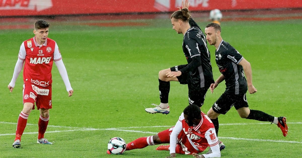 Fußballspieler in Aktion; ein Spieler springt, ein anderer rutscht und ein dritter fällt mit dem Ball. Das Feld ist nass und grün.