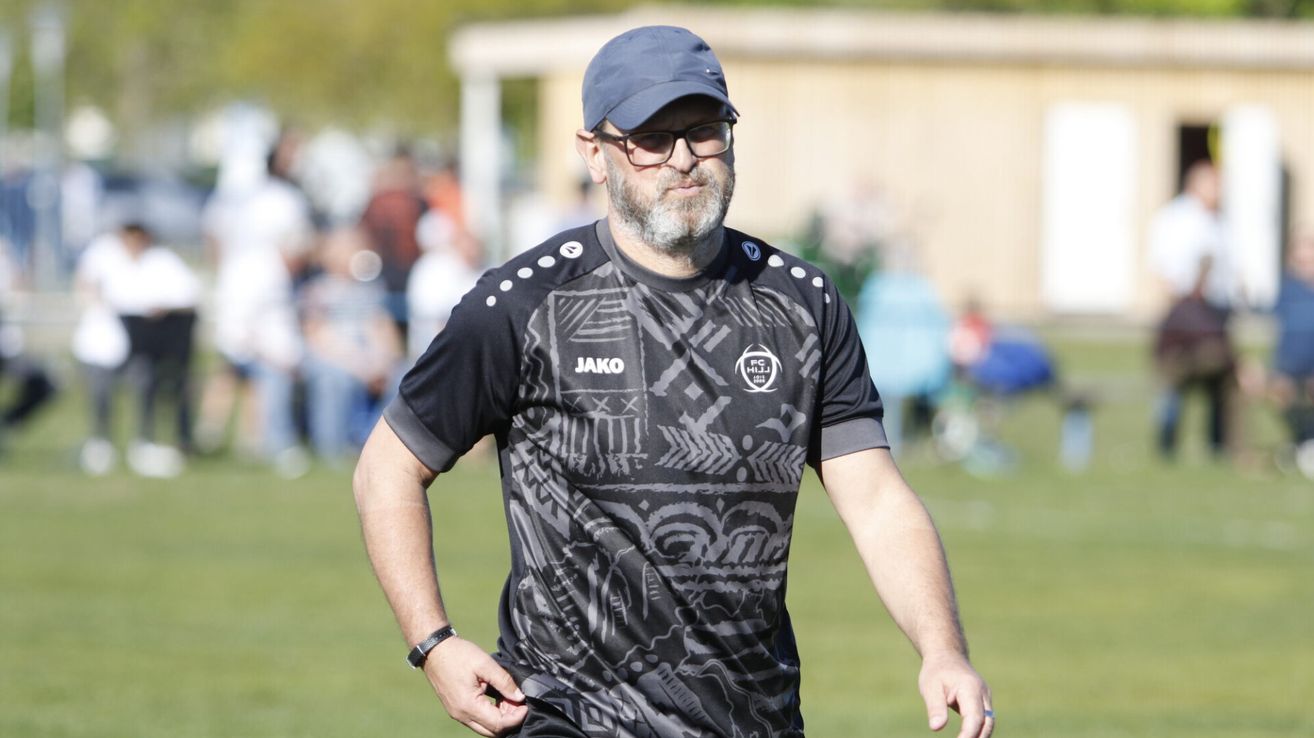 Ein Mann mit blauer Mütze und Brille geht auf einem Fußballfeld. Er hat einen Bart und trägt ein schwarzes Trikot mit weißen Designs.
