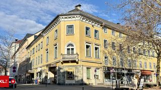 Ein städtisches Gebäude mit mehreren Fenstern und einem Balkon an der Ecke, mit einem klaren blauen Himmel darüber und Geschäften im Erdgeschoss.