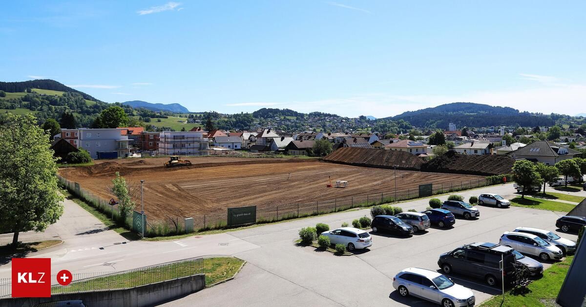 Luftaufnahme eines Baugeländes mit einem großen Feld, einem geparkten gelben Bagger und einigen Häusern. Autos sind auf der Straße und im Parkplatz geparkt.