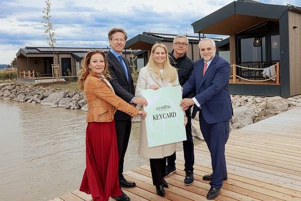Five individuals stand on a wooden dock, presenting a green placard reading 'KEYCARD'. Behind them, a serene lake and modern houses are visible.