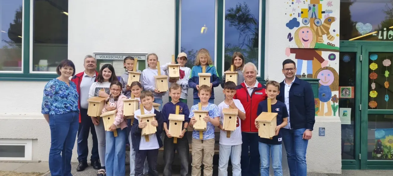 Eine Gruppe von Kindern und Erwachsenen steht draußen und hält hölzerne Vogelhäuschen. Die Kinder lächeln und scheinen für ein Foto zu posieren. Der Erwachsene im roten Mantel lächelt ebenfalls und steht in der Mitte. Hinter ihnen befindet sich ein Schild an einer Wand mit dem Wort 'Schule' darauf.