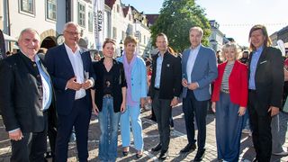 Eine Gruppe von Menschen in formeller Kleidung steht auf einer Kopfsteinpflasterstraße. Hinter ihnen befinden sich Gebäude mit Bannern.