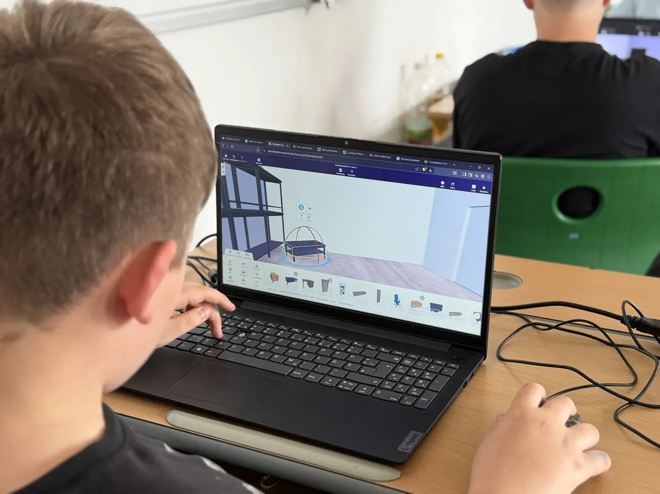 Ein Schüler benutzt einen Laptop, der ein 3D-Modell auf einem Schreibtisch anzeigt. Der Laptop-Bildschirm zeigt einen Raum mit Möbeln. Eine weitere Person sitzt im Hintergrund.