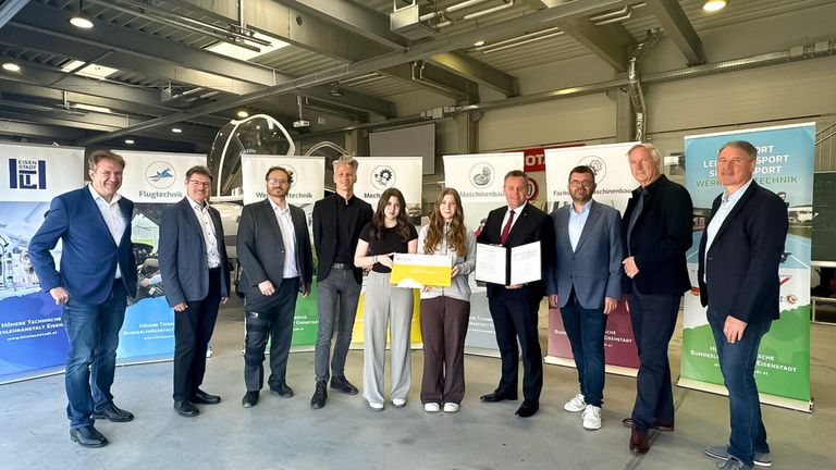 Mehrere Personen posieren mit einem Zertifikat in einem gut beleuchteten Raum mit Bannern und einem großen Glasobjekt.