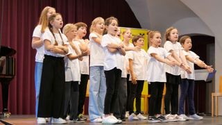 Eine Gruppe von Kindern, alle in übereinstimmenden weißen T-Shirts und schwarzen Hosen gekleidet, steht auf der Bühne und führt möglicherweise ein Lied oder einen Musiknummer auf.