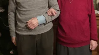 Zwei ältere Personen stehen vor einem geschmückten Weihnachtsbaum und lächeln für ein Foto.