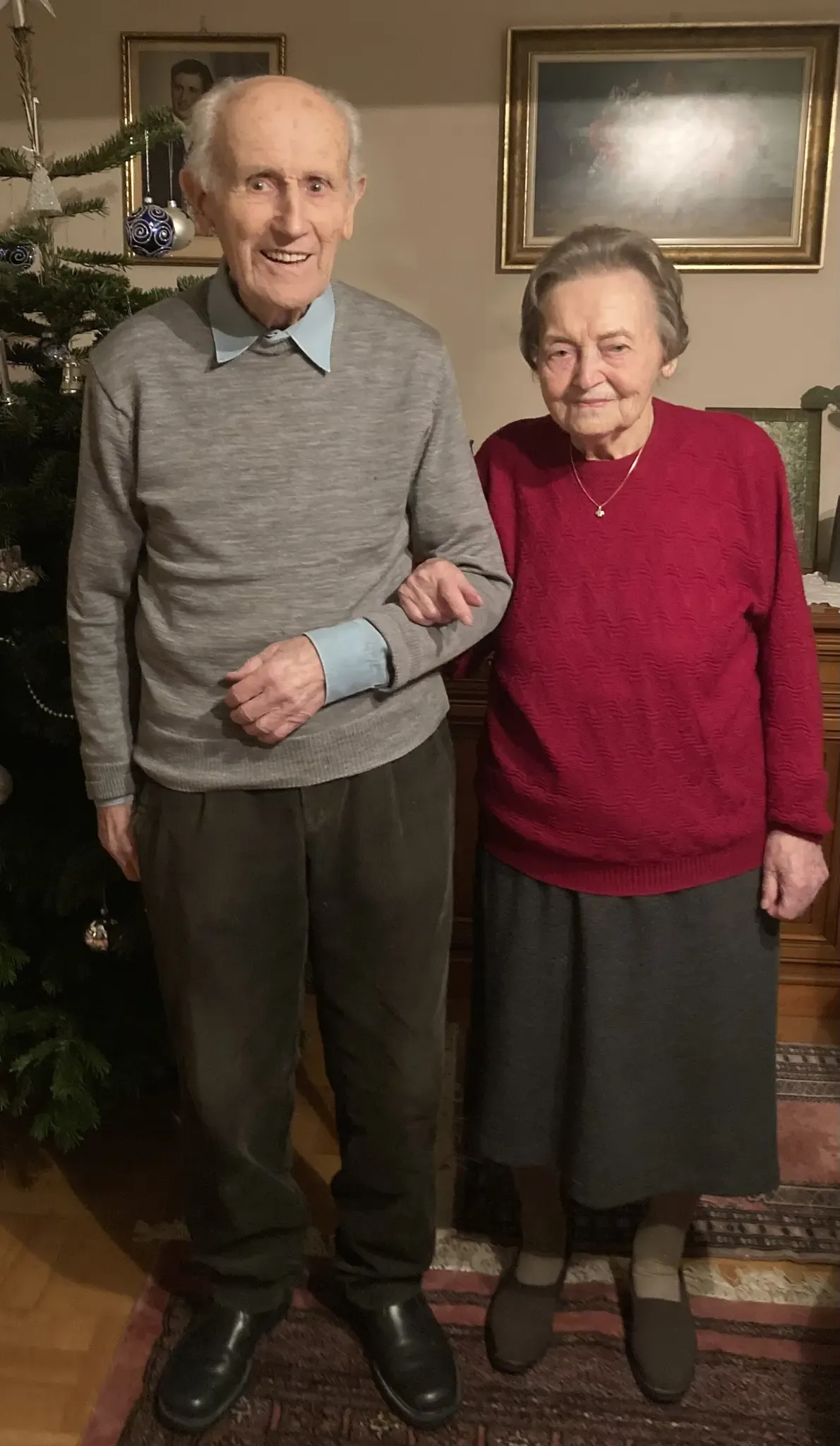Zwei ältere Personen stehen vor einem geschmückten Weihnachtsbaum und lächeln für ein Foto.